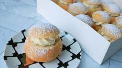 A semla bun on a plate next to a box full of semlor buns.