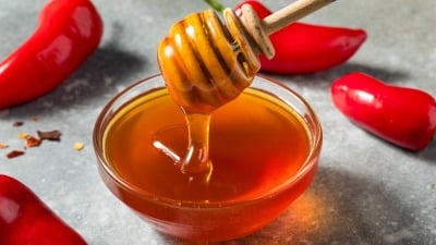 A honey dipper drizzling honey into a bowl with red peppers in the background.