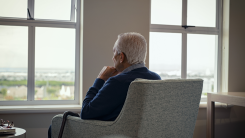 An elderly man in a care facility, sitting in a gray chair and gazing out a window